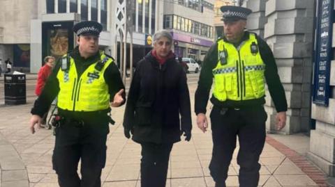 A woman with grey hair tied back in a black coat and black trousers is walking through Sheffield city centre with two male police officers either side of her