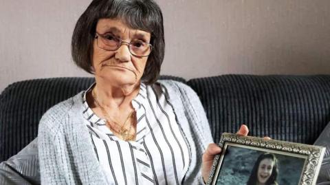 A woman with dark grey hair and glasses, white blouse and grey cardigan on a dark grey sofa holding up a picture of her daughter in a gold frame. She has long dark hair. 