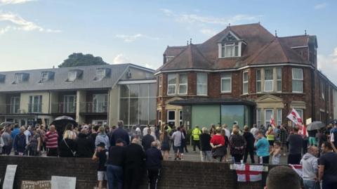 A group of people, some holding St George's flags, protesting outside a hotel.