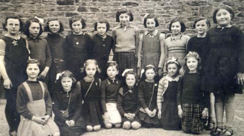 A black and white picture of evacuated children at school in Cockermouth during the Second World War. They are all girls arranged in a group posing for the picture. One row of children is standing in the back, with another row kneeling in front of them.