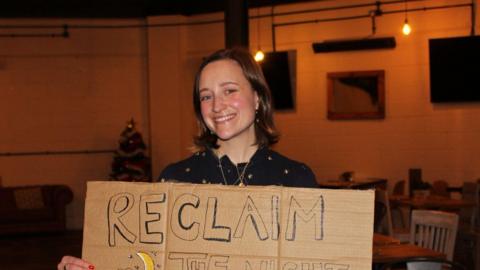 Woman with brown bob haircut smiles at camera, holding a sign which says 'Reclaim the Night, Reclaim These Streets'.