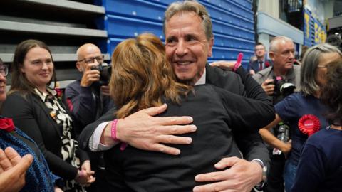 Dan Norris hugging a woman, surrounded by journalists and Labour supporters