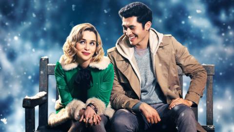 Emilia Clarke (as Katarina Andrich) and Henry Golding (as Tom) in the film Last Christmas. They are sitting on a bench and smiling, it is snowing in the background. 
