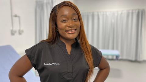 Dr Miranda Ntorinkansah smiling at the camera. She is wearing black medical scrubs which have her name embroidered on them.