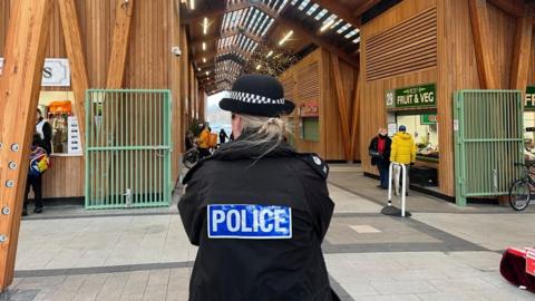 A police officer stands, looking, within Great Yarmouth's market building. It is a timber building, with vaulted and semi-glazed roof, paved flagstones and featuring green steel gates.