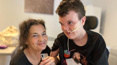 Sam, is a woman with brown wavy hair. She is wearing a black top, kneeling next to Alex's wheelchair smiling for the camera. Alex is a boy in a black T-shirt, with a feeding tube attached to his nose.
