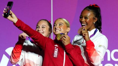 Image of three teenagers wearing tracksuits and holding up medals as they take a selfie standing on a podium.