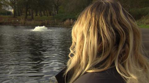 A woman who is a victim of crime who the BBC has agreed not to name, is looking away from the camera towards a water feature in a lake to avoid showing her face. She has blonde hair.