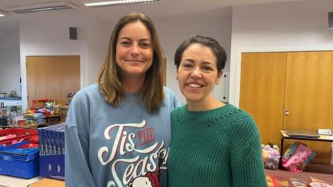 Two women stood side by side, looking into the camera and smiling. Laura, on the left, is wearing a blue jumper with 'Tis The Season' written on it in white letters. Penny, on the right, is wearing a dark green knitted jumper. They're pictured indoors, stood in front of rows of tables stacked high with crates full of food, toys, and toiletries.