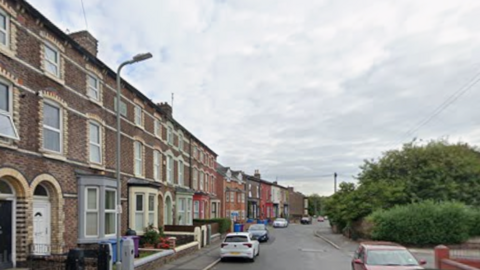 Image shows terraced houses on the left-hand side of Moscow Drive in Liverpool. Cars are parked along the road, and there are trees in a park on the right hand side of the photo. 