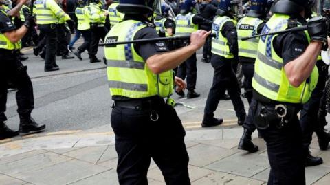 Police in Bolton town centre are armed with batons as they walk towards protesters. No faces are shown. 