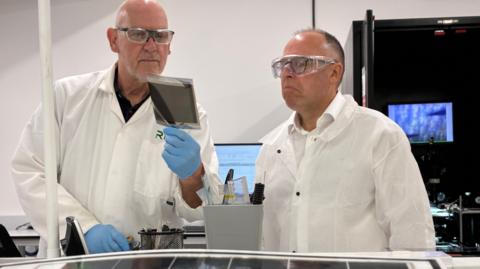 Two men in white coats and protective glasses look at a small black solar panel, with a sheet of small panels laid out on a table in front of them