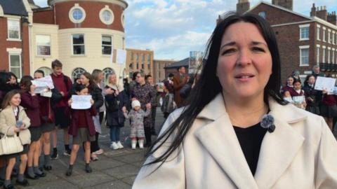 Anna Mulholland is wearing a cream coloured coat and a black top. She is standing in front of a group of children and parents outside LIPA School who are holding banners.