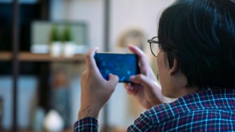 Back of the head of a man wearing glasses sitting down playing a video game on a mobile phone