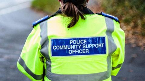 A female police community support officer stands with her back to the camera. She is wearing a yellow hi-vis jacket