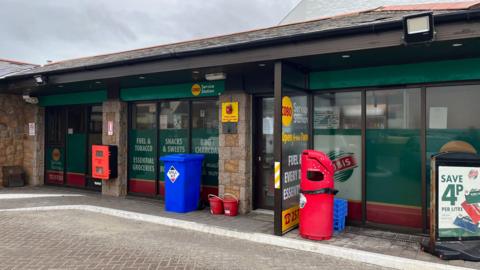 A picture of a garage. It is a brick and glass building. There is bins outside of it. The building is green and red in colour.