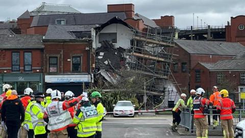 A partially collapsed building that shows roof tiles strewn over a pile of debris a scaffold hangs nearby. A number of emergency workers stand nearby.