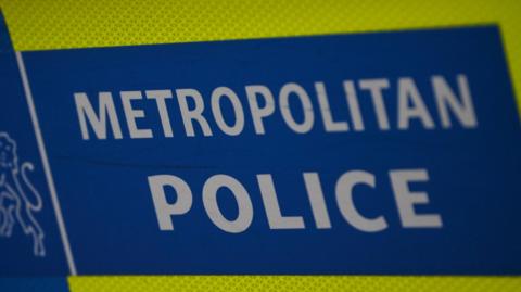 A close up Met police livery on a police vehicle. It is blue with white letters spelling Metropolitan Police with a yellow strip visible above and below. 