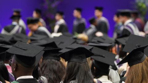 Rows of Degree students wearing gowns and black mortarboards. The background in the distance is purple.