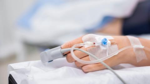 A hand with a surgical dressing and a monitoring clip on the index finger lies on a hospital bed.