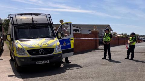 Three police officers stand next to crime scene tape and a police van. Behind them, a tarmac area with red brick wall, forensics tent and small cones.
Behind the wall, the tops of small dwellings can be seen.