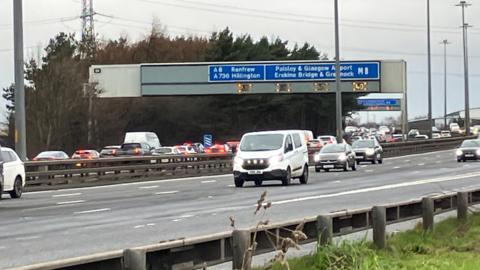 Section of M8 showing backed up traffic in the westbound lane, free flowing traffic in the eastbound lane and a sign overhead which reads: "A8 Renfrew, A736 Hillington, M8 Paisley & Glasgow Airport Erskine Bridge & Greenock"