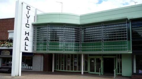 A flat roofed building with green windows and frontage and a large white horizontal sign attached to the left side of the building that says Civic Hall