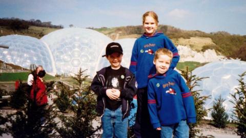 Rosie Johns and her two brothers are posing for the photo and behind them are the biomes. Rosie and one of her brothers are wearing Harry Potter branded hoodies and the other brother is wearing a cap. One of the brothers is pulling a funny face.