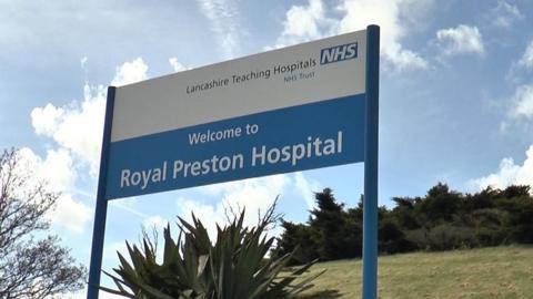 A blue and white sign reading 'Welcome to Royal Preston Hospital' in front of green bushes and and blue cloudy sky