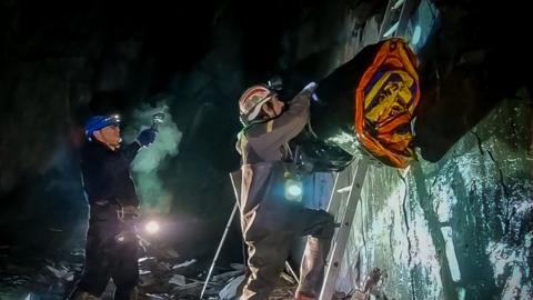 Volunteer Gerard Carton lifts a roll of several deflated dinghies above his head as he climbs up a ladder from a mine shaft, with another volunteer below shining a light on him