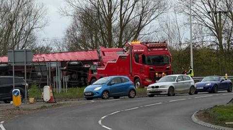 A red lorry broken down on the edge of a roundabout. Two men can be seen standing in the road at the front of the lorry looking at it. In front of the roundabout are queuing cars. 