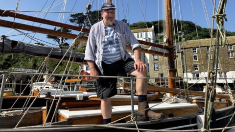 There is a man smiling on the deck of a yacht. He has a baseball cap over a bandanna. He is wearing different check tops, black shorts and socks inside boots. Behind him is the harbour walls and properties