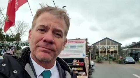 Michael Moody standing in the car park outside Booths supermarket. He is wearing a coat, a white shirt and green tie. Behind him is a billboard explaining his protests and a set of household shelves.