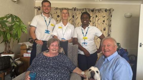 A woman in a flowery blue top and a man in a blue shirt, with a dog and three people in doctors uniform.