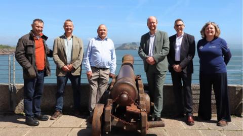 Six people stood next to a cannon (three either side). In the background is the sea and a small island (Drakes Island). The sky is blue. 