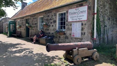 An old stone building with a sign saying Sark Visitor Centre: La Societe Serquaise Heritage Room (meaning The Sark Society in Sark's local language). There is a canon in front, hanging baskets, a green telephone box and vines running up the side wall. A woman sits on a bench outside.