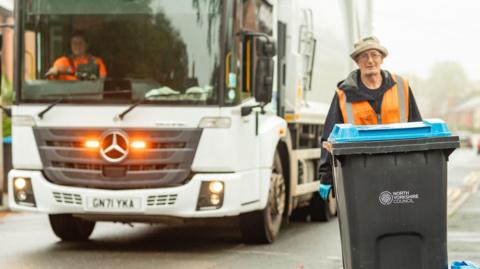 Man with an orange hi-vis jacket wheeling a bin in front of a lorry