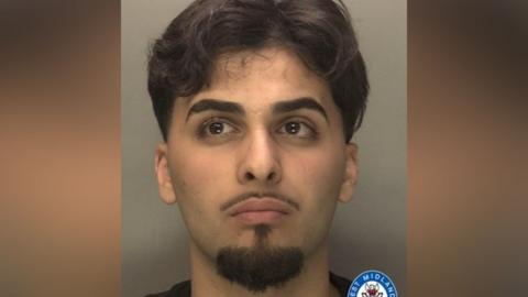 Deara Sadiq has long black hair and black beard, goatee and moustache. He is wearing a black top.