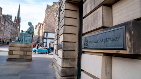 A general view of the sign at the entrance to the high court in Edinburgh. A black sign is attached to a light-stone brick wall. It reads High Court of Justiciary'.
