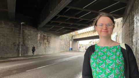 A woman with a bright green pigeon dress and glasses standing by the road under a bridge