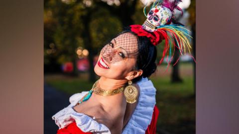 Red fascinator with skull with colourful illustrations. She is wearing an oufit with white frils, red body and a yellow/pink/yellow dress