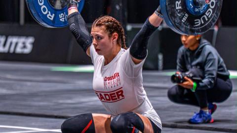 A teenager with long dark, braided hair and wearing a white shirt, shorts and knee pads, crouches down as she lifts a bar with weights on either end. 