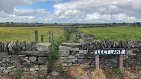 A road sign with Fleet Lane on it in black lettering in front of a drystone wall and green fields with yellow flowers in the distance. 