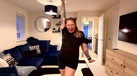 A woman in a black t-shirt and shorts, with glasses and long dark blonde hair. She has her arms outstretched and she s smiling. She has several tattoos on her arms. She is in a living room which has a blue sofa, tv and mirror on the wall. 