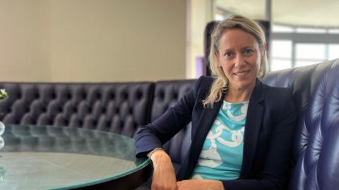Alice Armanni Sequi is sitting on a dark blue tufted leather couch in a modern, well-lit room. She is wearing a light blue dress with white patterns and a dark blazer. There is a round glass table in front of the person with some small objects on it. The background shows large windows and part of another room
