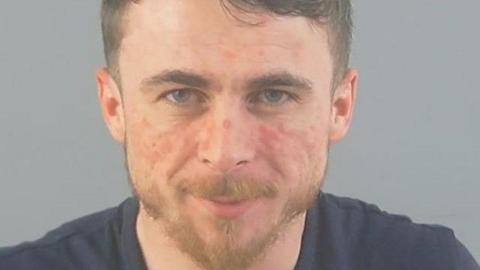 Scott John William Smith, a man with reddish marks on his face and ginger facial hair, appears to smile slightly at the camera in a police mugshot.
