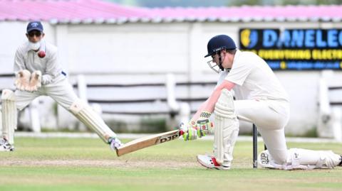 A vicar drives a ball with a wicket-keeping imam in the background