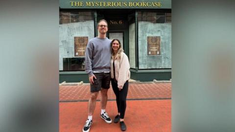 Ally Fir (R) and Matt Rawlings (L) pictured outside their shop before it opened. The windows are empty and there is a sign saying "The Mysterious Bookcase". 