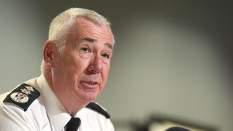 PSNI chief constable Jon Boutcher, with short grey hair, pictured from the shoulders up. He is to the left of the image. He is wearing a white shirt and black tie. 