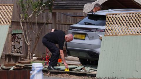 A man leans down to place a yellow marker of evidence on the ground amid rubble and fencing after a silver car beside him crashed through a fence leading to a nursery playground.